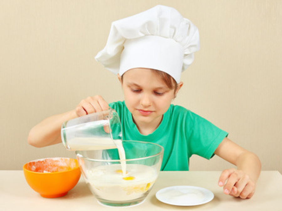 Kind mit Kochmütze auf Kopf gießt Milch in Schüssel